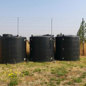 Three black water tanks in a dry, grassy field with a wire fence.